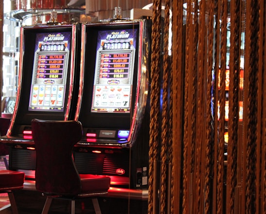 A pair of casino slot machines, each with detailed screens displaying various game outcomes and jackpot amounts. The chairs in front of the slot machines are dark and cushioned for comfort. The background features a hanging curtain made of golden ropes and other casino decorations are visible.